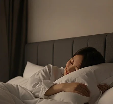 침대에 누워 있는 한 사람이 보이며, 파란색 벽과 베이지색 헤드보드가 있는 침실의 모습입니다. 사람은 흰색 잠옷을 입고 흰색 베개와 이불에 둘러싸여 있습니다. 방 안은 조용하고 편안한 분위기를 자아내며, 조명이 은은하게 비추고 있습니다.