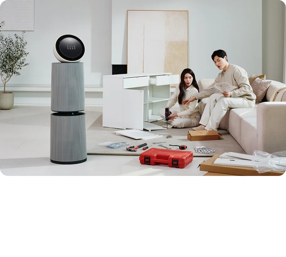 거실 공간에 LG 퓨리케어 AI 오브제컬렉션 360˚ 공기청정기가 놓여 있습니다. 가구를 조립하고 있는 두 사람의 모습이 보입니다. 주변에는 조립 중인 서랍장과 공구가 있습니다.