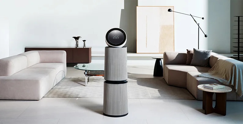 거실 공간에 LG 퓨리케어 AI 오브제컬렉션 360˚ 공기청정기가 놓여 있습니다. 양쪽에 베이지색 소파가 있으며, 쿠션과 담요가 놓여 있습니다. 유리 상판의 원형 테이블이 있고 그림과 검은색 스탠드 조명이 보입니다.